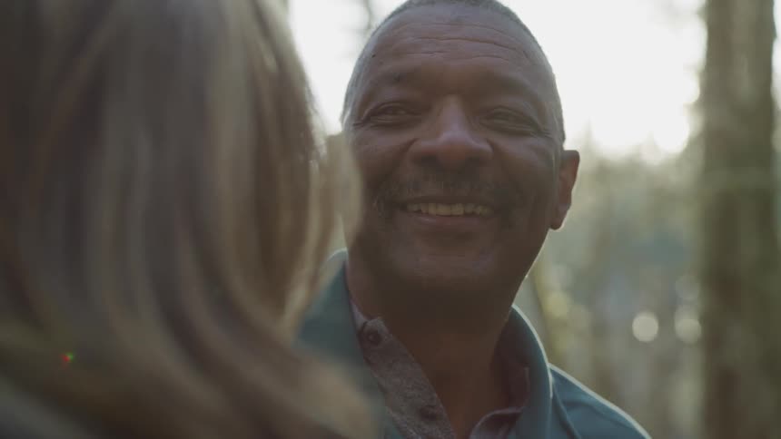 A man smiles while looking at a woman outside near trees