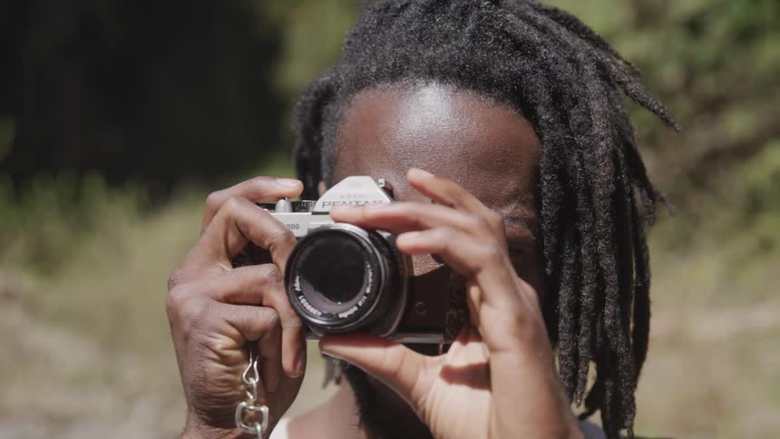 Black man with dreadlocks takes a picture with a camera in the forest