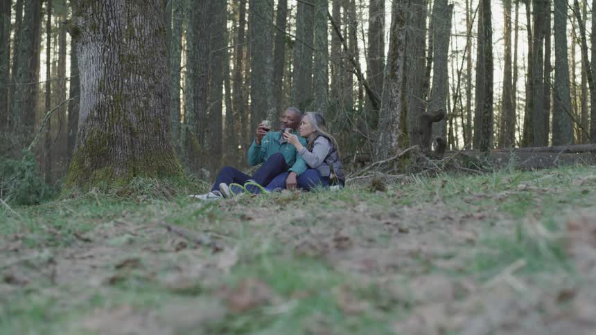 An older couple is sitting on the ground in a forest drinking red wine from wine glasses