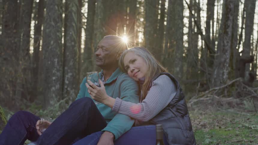 A woman holds a glass while she sits with her head on her husbands shoulder near the trees