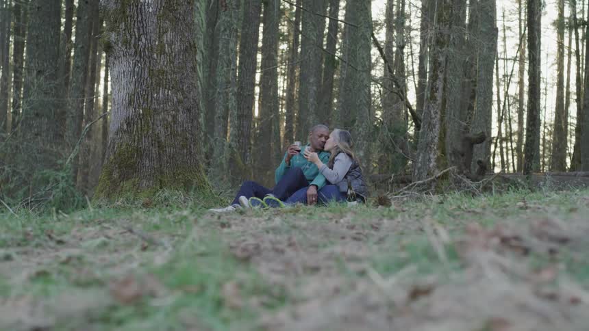 A man and woman kiss while drinking wine near forest trees