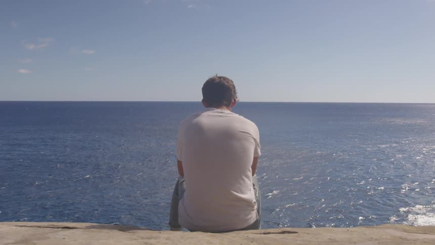 A man sits on a beach and looks out towards the water