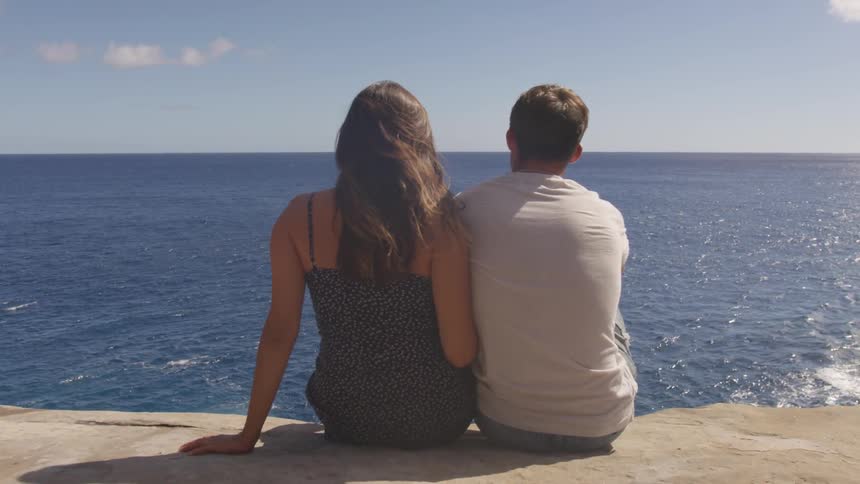 A man and woman sit on a cliff and look out at the ocean