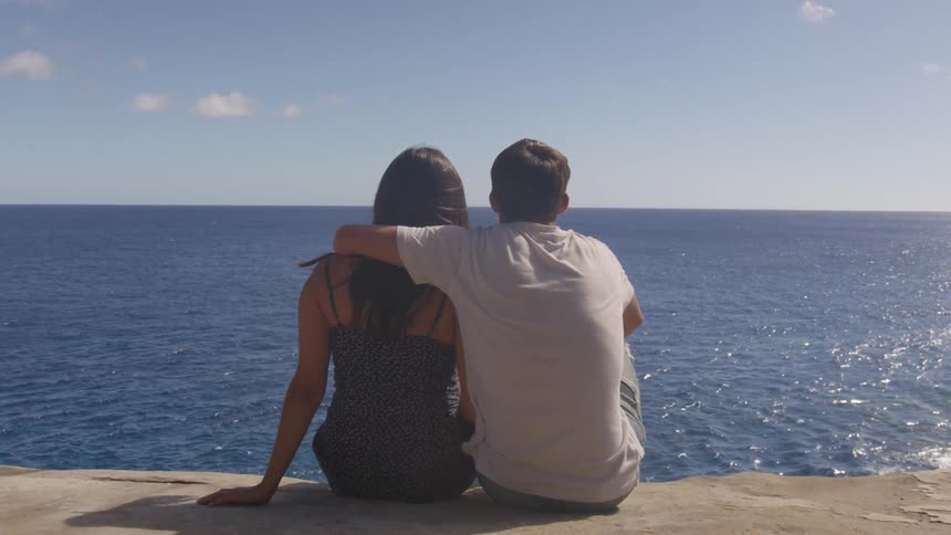 A young couple sit on the edge of a cliff and look out over the ocean