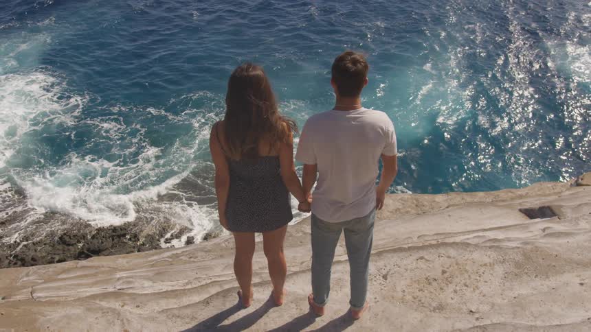 Couple holds hands standing on cliff over ocean