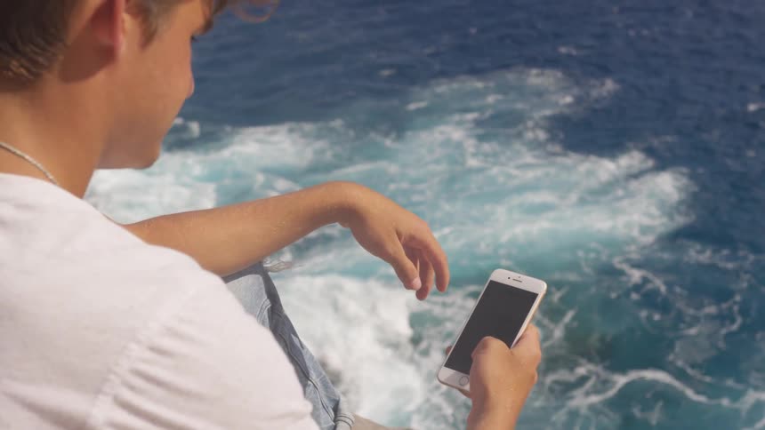 A young man uses his cell phone while sitting above the ocean