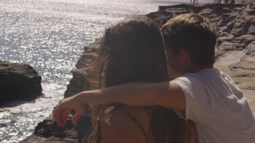A boy hugging a girl as they sit down and watch the ocean