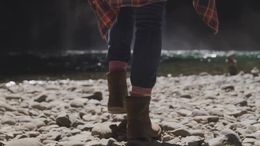 A woman wearing hiking boots walks on rocks toward the edge of a river