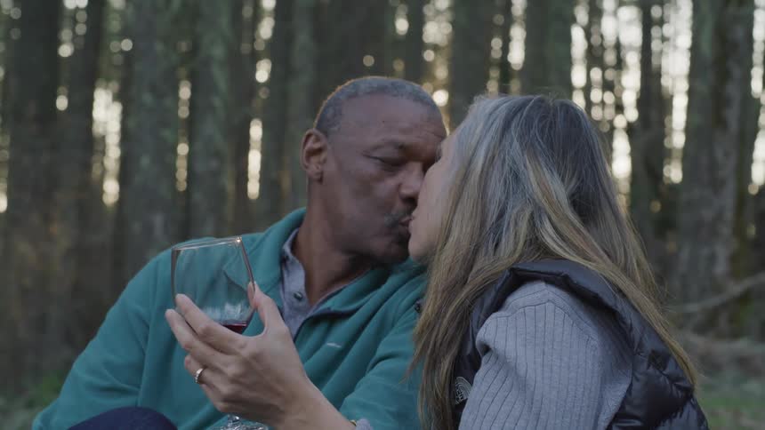 Black man and blonde woman holding a glass of wine kiss each other in the forest at sunset