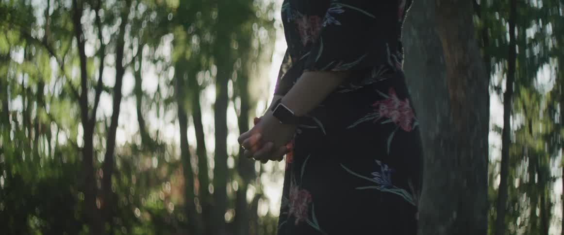 Woman clasping hands stands on the grass while the sun streams through the trees