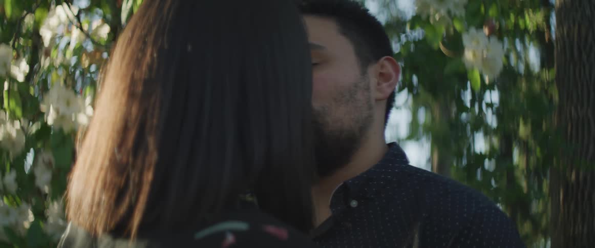 A man leans forward and kisses his girlfriend outside near flowering trees