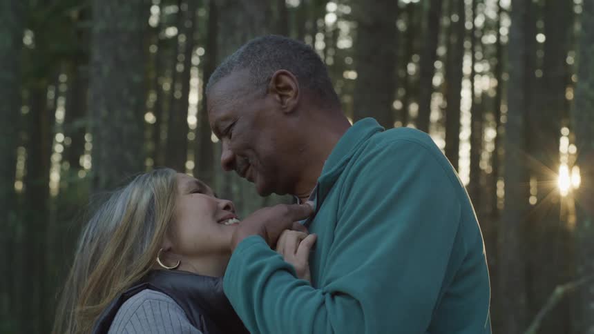 Black man and blonde woman hold each other while the sun goes down in the forest