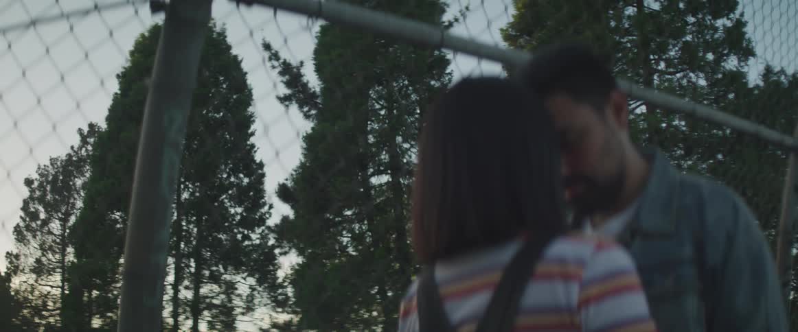 Man and woman stand close together in front of a chain link fence