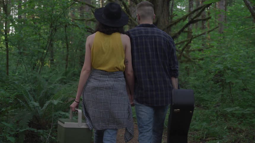 A white couple walk on a path through the woods while carrying a cooler and a guitar case