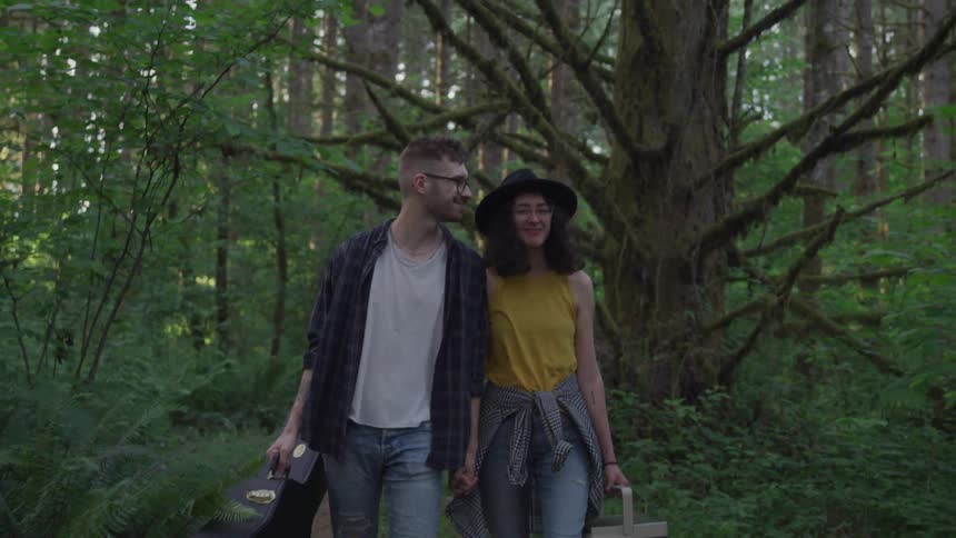 A man and woman hold hands while carrying a guitar case and cooler in a forest