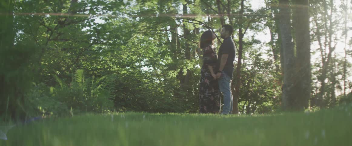 Young couple holding hands outside on a grassy area near some bushes and trees