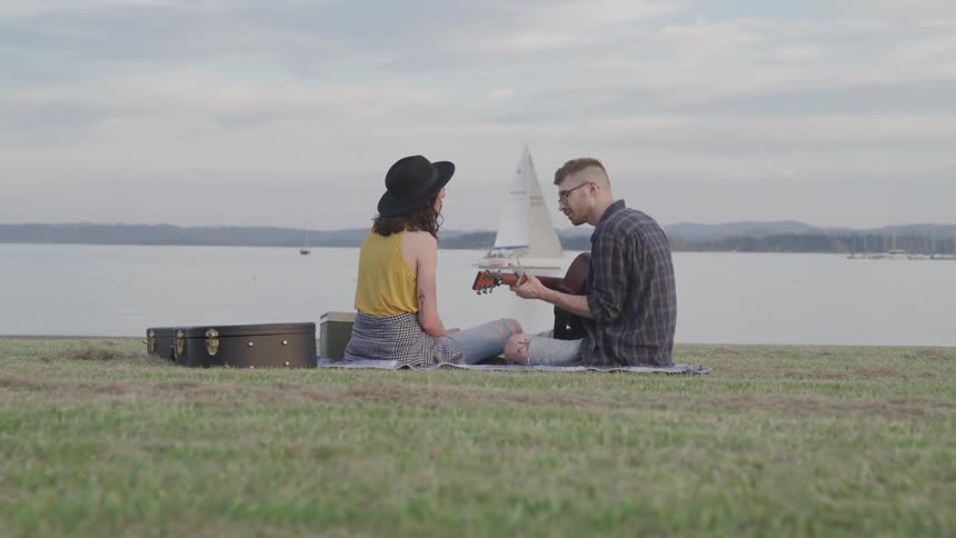 Woman and man who plays a guitar sit on the grass next to the sea