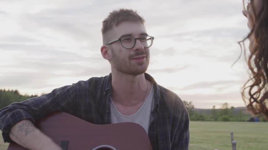 Man and woman sitting on the grass play the guitar and sing a song at sunset