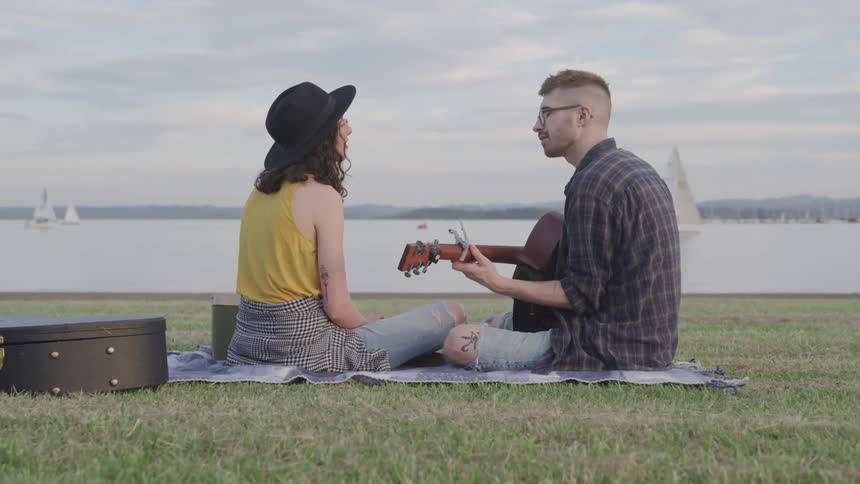 A man plays the guitar for his girlfriend as they sit on a blanket in front of the lake