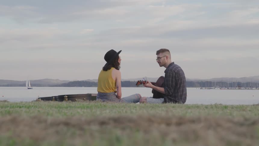 A man and woman sit beside a lake and the man plays the guitar