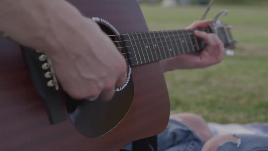 Person strums guitar sitting outdoors on a blanket