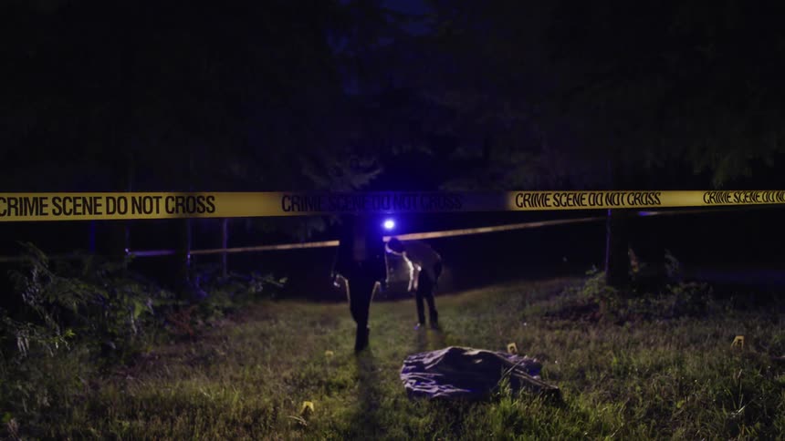 Two police officers approach a covered body surrounded by police tape at night