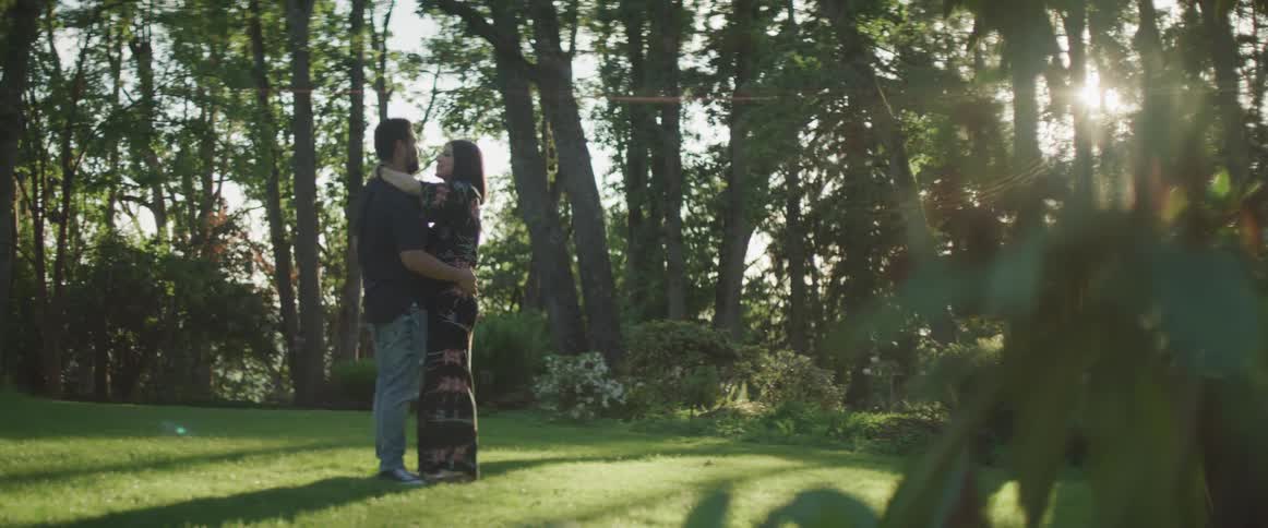 A man and woman slow dance on the grass clearing surrounded by trees on a sunny day