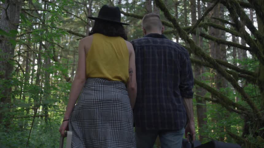 A woman carries a picnic basket and a man carries a guitar while holding hands and walking through a forest