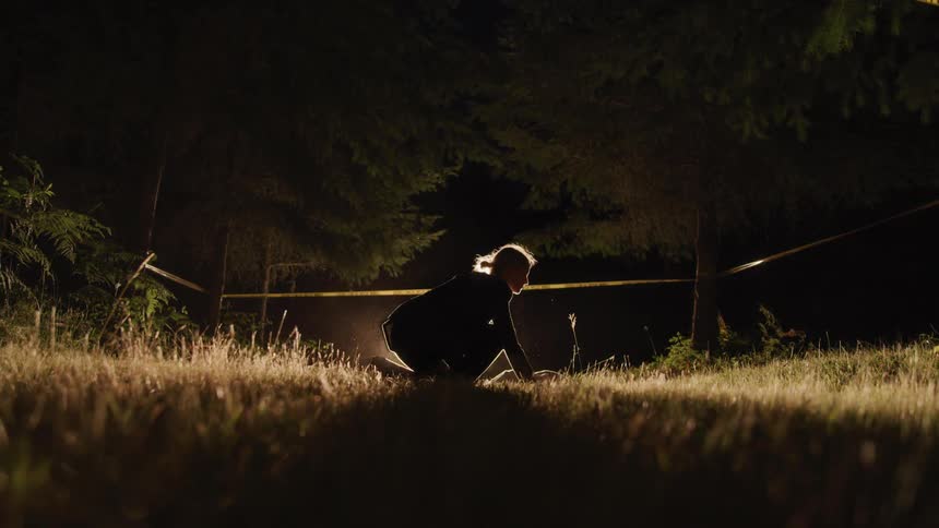 A young woman bends down behind some caution tape and examines a body on the ground