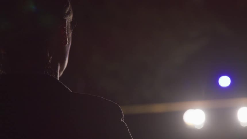The silhouette of a woman can be seen illuminated by the lights of a nearby police car at a nighttime crime scene