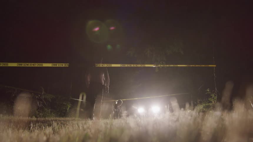 Man crosses crime scene tape surrounding area lit by car headlights