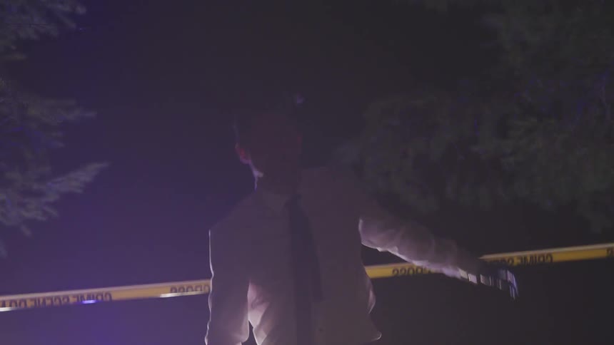 A man with a shirt and a tie and disposable gloves ducking under a crime scene tape then walking and squatting