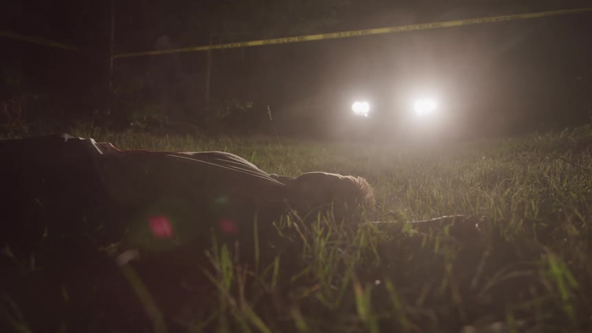 A young man lies in a field with headlights shining on him and caution tape bordering the area