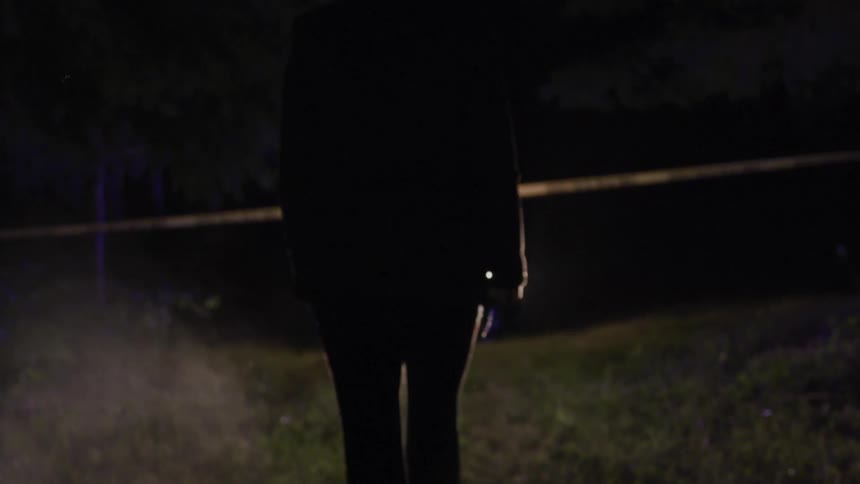 A person in a police cordon located in a field walking towards a vehicle with its headlights on