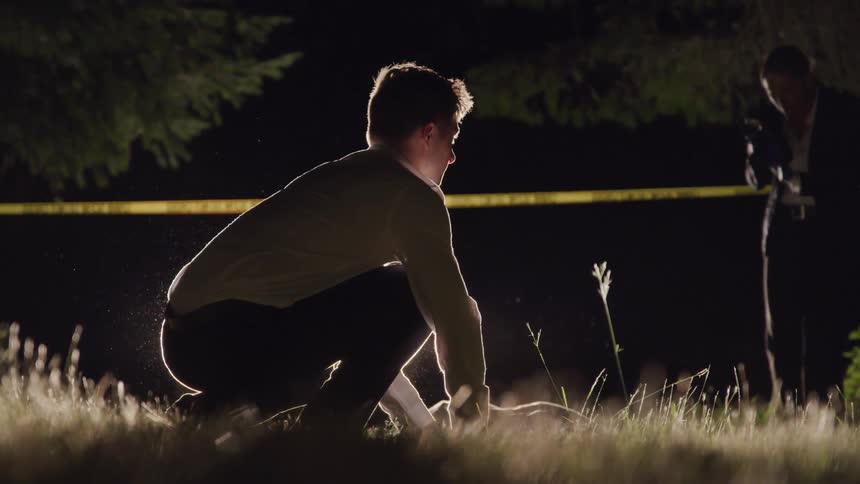 A detective kneeling to examine a corpse as a forensic photographer takes photos