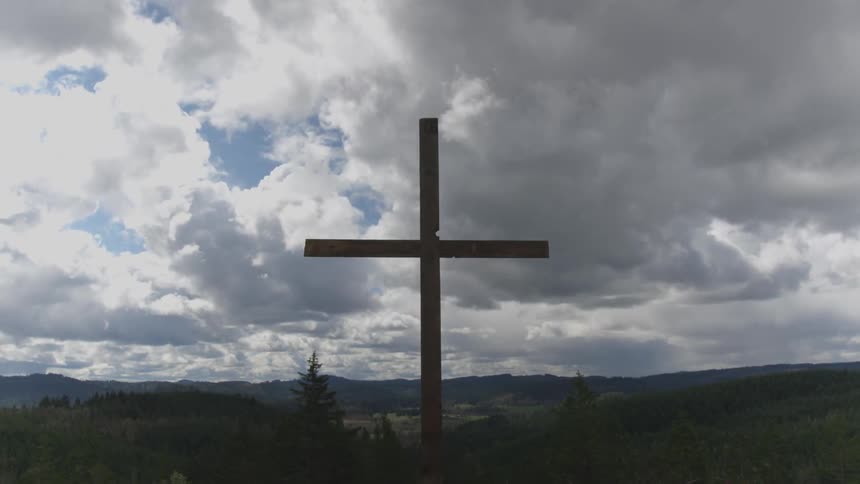 A tall wooden cross stands in front of a forest as clouds pass quickly overhead