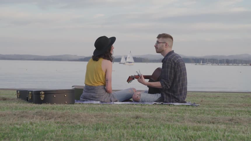 A man plays guitar and sings to a woman while sitting on a blanket on a lawn