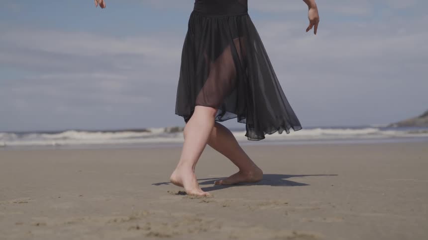 A woman points her toes and and flexes as she dances barefoot on the sand of an empty beach