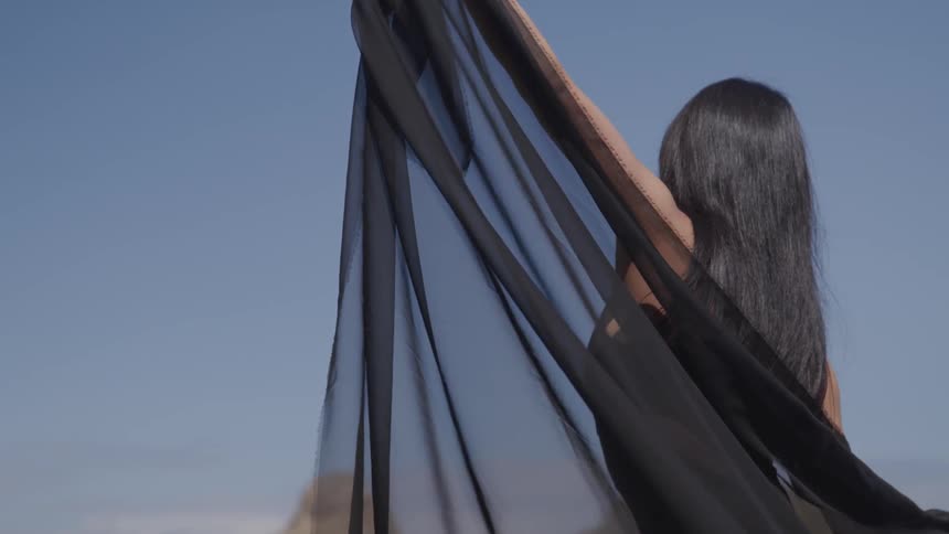 A woman holds up a sheer black fabric shawl with her arms stretched wide on a sunny blue sky day