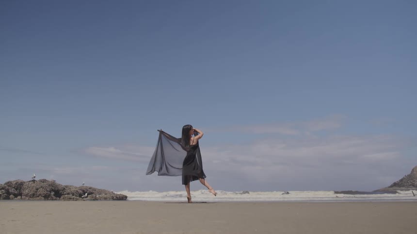 A woman walks along a beach with a cape flowing behind her