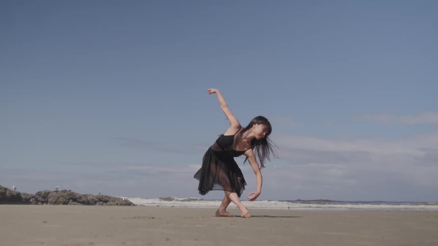Woman in black dress dancing ballet on the sand of beach next to the ocean