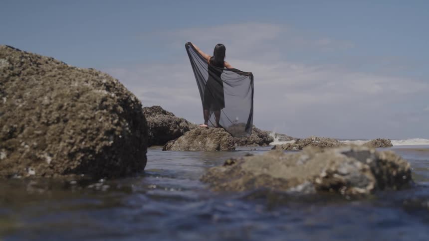 A woman in a shawl dances on rocky coastline