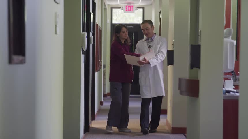 A doctor walks down a hallway with a woman who points at the open folder he is carrying