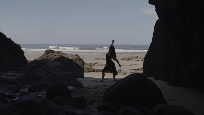 Asian womans silhouette in black dress dances on the sand beach between big rocks