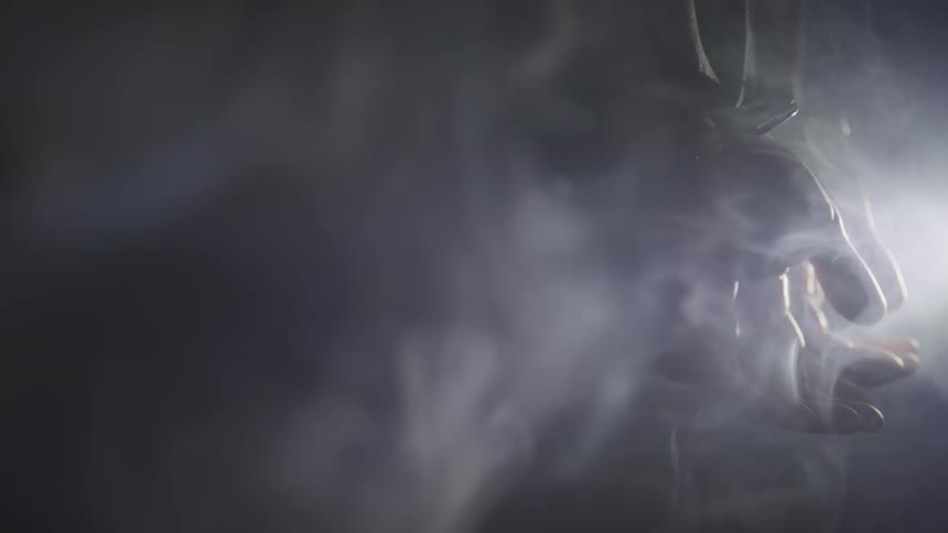 A man stands in darkness with his hands cuffed behind his back as smoke rises around him