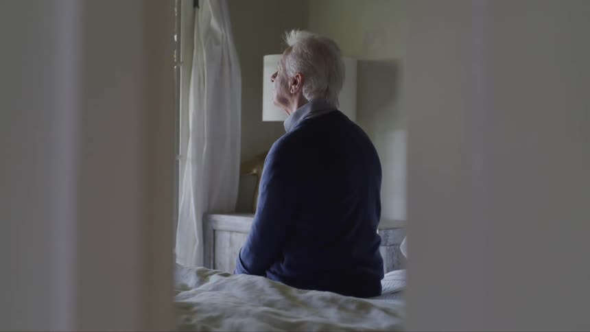 An elderly man sits on a bed and looks out the window