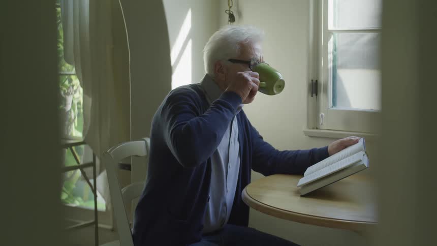An elderly man sits at a wooden table reading a book and drinking out of a mug