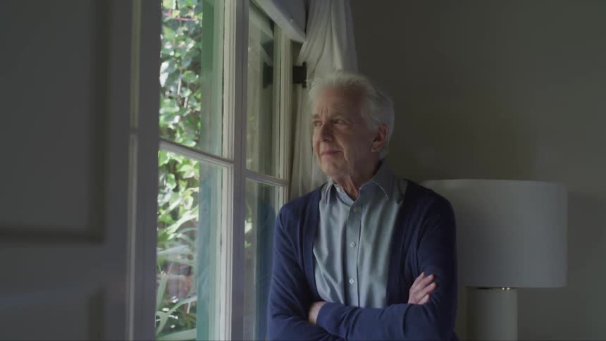 An elderly man with white hair stands with his arms crossed in front of him while he looks out a window
