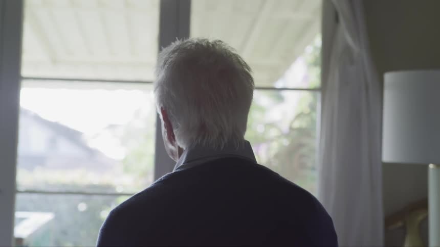 An elderly man in a collared shirt and sweater is staring out a window