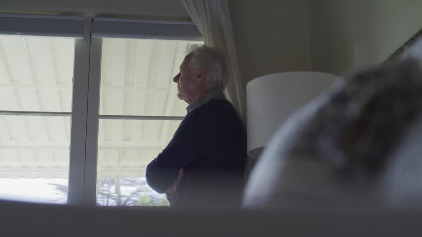An elderly man with white hair stands with his arms crossed while looking out the window